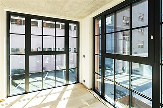 Světlý obývací pokoj s velkými okny, parketovou podlahou a moderním designem, umožňující přirozené osvětlení a výhled do vnitrobloku