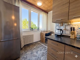 Kuchyň s moderními dřevěnými skříňkami, černou pracovní deskou, velkými okny a dlažbou, nabízí světlý a funkční prostor pro vaření.