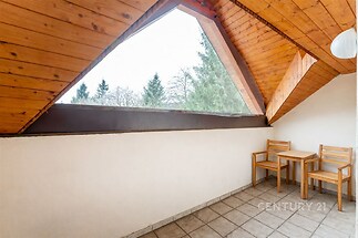 Podkrovní terasa s dlažbou, dřevěným obložením stropu a velkým štítovým oknem nabízí klidné posezení s výhledem na stromy.