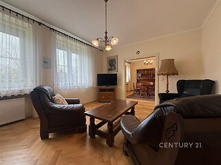 Světlý obývací pokoj s parketovou podlahou, velkými okny a koženým nábytkem, propojený s jídelnou, vytváří útulný a elegantní prostor.