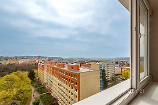 Výhled z okna na panoramatické město, moderní okolí, panelové domy s barevnými fasádami, svěží zeleň v okolí.