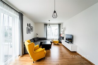 Světlý obývací pokoj s parketovou podlahou, velká okna a moderní vybavení. Prostor je elegantní a stylově zařízený s barevnými akcenty.