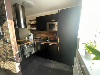 Moderní kuchyň s černými dvířky, dřevěným obložením a dlažbou. Vestavěné spotřebiče a velká okna zajišťují světlý interiér.