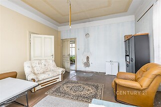 Světlý obývací pokoj s vysokým stropem, dřevěnou podlahou a velkým oknem. V místnosti je potřeba rekonstrukce.