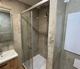 Moderní koupelna s keramickými obklady, proskleným sprchovým koutem a dřevěnou skříňkou pod umyvadlem, vše v rekonstruovaném stavu.