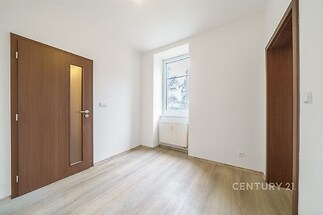 Světlý pokoj s parketovou podlahou, velkým oknem a moderními dveřmi. Bílá stěna a přírodní dřevěný design podtrhují elegantní styl.