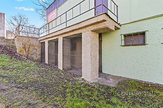 Přiléhající prostor s betonovými sloupy a hrubou omítkou, vhodný pro úpravy, vedle zatravněného pozemku, ideální pro skladování nebo dílnu.