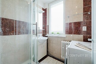 Koupelna s moderní dlažbou v kombinaci hnědé a béžové barvy, velké okno pro dostatek světla, prostorný sprchový kout a pračka.