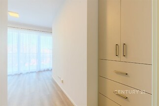 Chodba s vestavěnými skříněmi a světlou parketovou podlahou, vedoucí do místnosti s velkými okny a moderními žaluziemi.