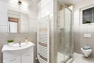 Moderní koupelna, světlé dlažby, sprchový kout s prosklenými dveřmi, nástěnný radiátor, čistý design, malé okno s květinou.