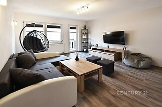 Světlý obývací pokoj s moderním nábytkem, parketovou podlahou a velkými okny. Elegantní závěsné křeslo a minimalistický design dodávají prostoru styl.