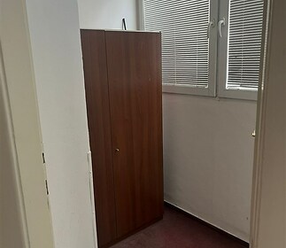 Malý pokoj s kobercem a vysokou dřevěnou skříní, která se nachází pod oknem se žaluziemi, ideální jako úložný prostor nebo pracovna.