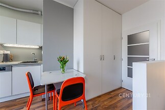 Kuchyňský kout s moderními bílými skříňkami, parketovou podlahou a světlým jídelním stolem s barevnými židlemi.