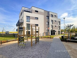 Moderní bytový dům s balkonem a dětským hřištěm před budovou, oplocená zahrada, novostavba v klidné lokalitě.