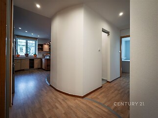 Prostorná chodba s moderní vinylovou podlahou, zaoblené stěny vedou do kuchyně s velkými okny a vestavěnými spotřebiči.