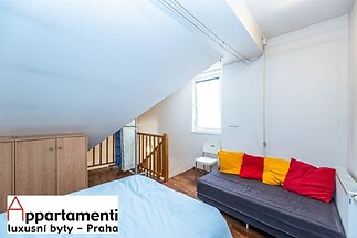 Podkrovní ložnice se dřevěnou podlahou, šatní skříní a světlým oknem, moderní interiér s barevnými polštáři na rozkládací pohovce.