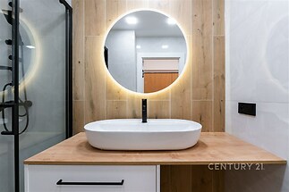 Moderní koupelna s dřevěným obkladem, umyvadlem na desce a LED osvětlením. Skleněný sprchový kout a elegantní černé doplňky.