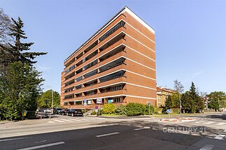 Červená cihlová fasáda bytového domu s velkými okny obklopený zelení v klidné rezidenční oblasti.