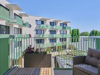 Prostorný balkon s výhledem na moderní bytový komplex, ratanový nábytek, zelené zábradlí, svěží venkovní posezení, slunečný den.