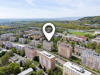 Pohled na sídliště s panelovými domy, obklopené zelení a přírodou. Budovy ve městě nabízejí klidné a dostupné bydlení.