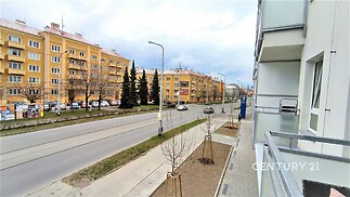 Balkon s moderním zábradlím s výhledem na městskou ulici s cihlovými domy, nedávno rekonstruované okolí s novými chodníky a zelení.