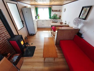 Kompaktní obývací pokoj s dřevěnou podlahou a krbovými kamny, propojený s kuchyní vybavenou moderními spotřebiči, světlý díky velkým oknům.