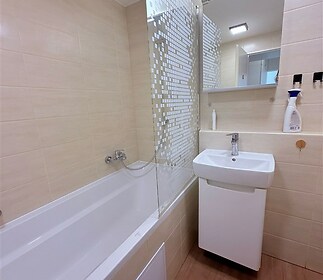 Moderní koupelna s dlažbou, vanou, a skleněným sprchovým panelem. Světlý interiér s úložnou skříňkou pod umyvadlem.