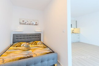 Kompaktní ložnice s moderní šedou postelí, světlými stěnami a světlou laminátovou podlahou sousedící s otevřenou kuchyní.