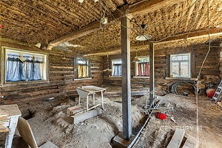 Rozestavěná místnost v rustikálním stylu, dřevěné trámy, hliněná podlaha, přírodní dřevo a velká okna.