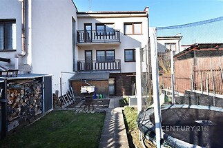 Zahrada rodinného domu s trampolínou a balkony. Dům má moderní omítku a dřevěné ohrady. Prostor pro uložení dřeva a venkovní posezení. Předzahrádka s chodníkem.