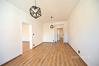 Světlý obývací pokoj s parketovou podlahou, moderním osvětlením a čistými bílými stěnami, vytváří elegantní a prostorný interiér.