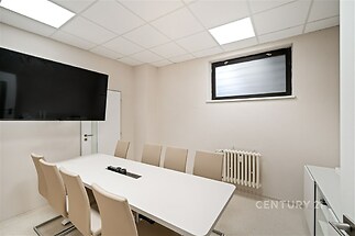 Moderní zasedací místnost s velkým stolem, ergonomickými židlemi a velkou obrazovkou. Světlý interiér s bílou dlažbou a minimalistickým designem