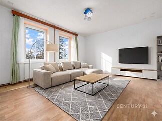Světlý obývací pokoj s parketovou podlahou, velká okna poskytující dostatek přirozeného světla, moderní nábytek a elegantní dekorace.