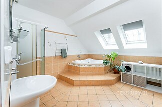 Podkrovní koupelna s dlažbou, rohovou vanou a dvěma střešními okny, která zajišťují dostatek světla. Moderní styl a praktické uspořádání.