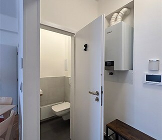 Vstupní předsíň s moderní dlažbou, světlé stěny a plynový kotel na stěně, vedle dveří vede úzká chodba k malé koupelně.