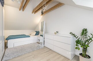 Podkrovní ložnice s dřevěnými trámy, moderním designem a parketovou podlahou, doplněná vestavěnými skříněmi a světlými stěnami.