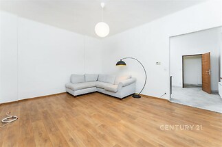 Světlý obývací pokoj s parketovou podlahou, moderní osvětlení a prostorný gauč, ideální pro pohodlné bydlení.