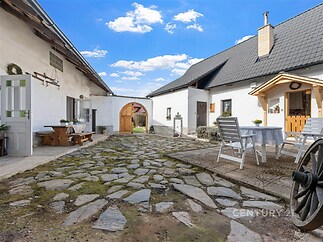 Venkovní dvůr s kamennou dlažbou a dřevěným nábytkem, rustikální styl, bílá fasáda budov, ideální pro relaxaci.