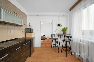 Kuchyňský kout s dřevěnou podlahou a moderními skříňkami, světlý prostor s velkými okny a jídelním stolem, ideální pro pohodlné bydlení.