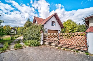 Rodinný dům s červenou střešní krytinou, dřevěným plotem a dlážděným vjezdem, obklopený zahradou, nacházející se v klidné lokalitě.
