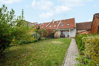 Zahrada s udržovaným trávníkem a dlážděným chodníkem, obklopena zelení. Klidné prostředí, vhodné pro rodinné posezení či rekreaci.