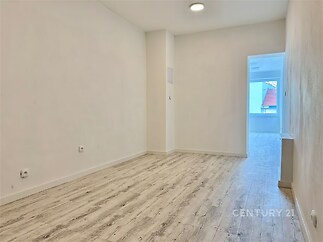 Prostorný pokoj s dřevěnou podlahou, světlými stěnami a moderními stropními svítidly, vedoucí do prosklené lodžie