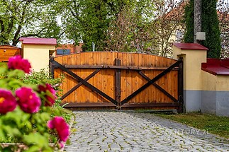 Dřevěná brána se nachází u vjezdu na dlážděnou příjezdovou cestu, obklopena kvetoucí zahradou a živým plotem v příjemném venkovním prostředí.
