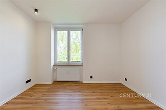 Prázdná místnost s parketovou podlahou, velkým oknem a moderním designem. Světlý interiér vytváří pocit prostoru a elegance.