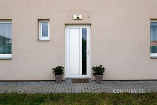 Vstupní dveře s moderním designem, obklopené dlažbou, dvorek s udržovaným trávníkem, světlá fasáda domu s okny pro přirozené osvětlení.