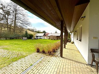 Prostorná terasa s dlážděnou podlahou nabízí výhled na rozlehlou zahradu, v pozadí se nachází přístavky a obklopuje ji přírodní zeleň