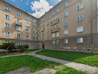 Venkovní pohled na bytový dům, plochá střecha, cihlová fasáda, rekonstruovaná okna, společný zatravněný dvůr, klidná lokalita.