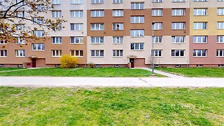 Panelový bytový dům s barevnou fasádou, před kterým je udržovaný trávník a příjezdová cesta, nachází se v klidné městské části.