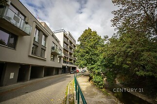 Moderní bytový dům s balkony, vedle zurčící potok, obklopený zelení, nabízí pohodlné a klidné bydlení ve městě.