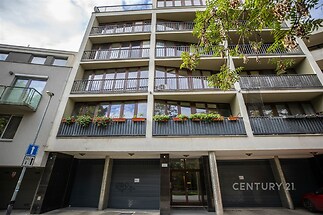Moderní bytový dům s velkými okny a balkony, nachází se na klidné ulici. Fasáda je betonová s kovovými doplňky, ideální pro městské bydlení.